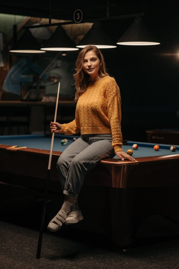 pexels-photo-7404679-7404679 Young woman in casual knitwear poses smiling on a billiard table indoors. Warm, relaxed atmosphere.